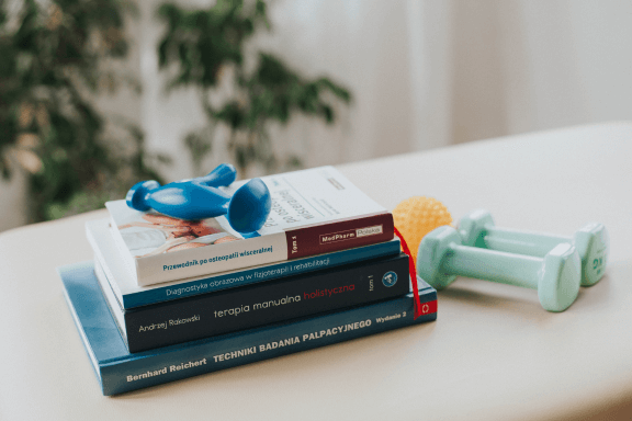 książki o fizjoterapii, hantle i piłka ułożone na łóżku do fizjoterapii