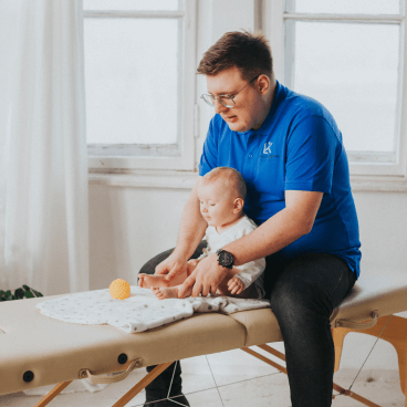 Fizjoterapeuta Łukasz Kusowski ćwiczący z siedzącym niemowlakiem na łóżku do fizjoterapii